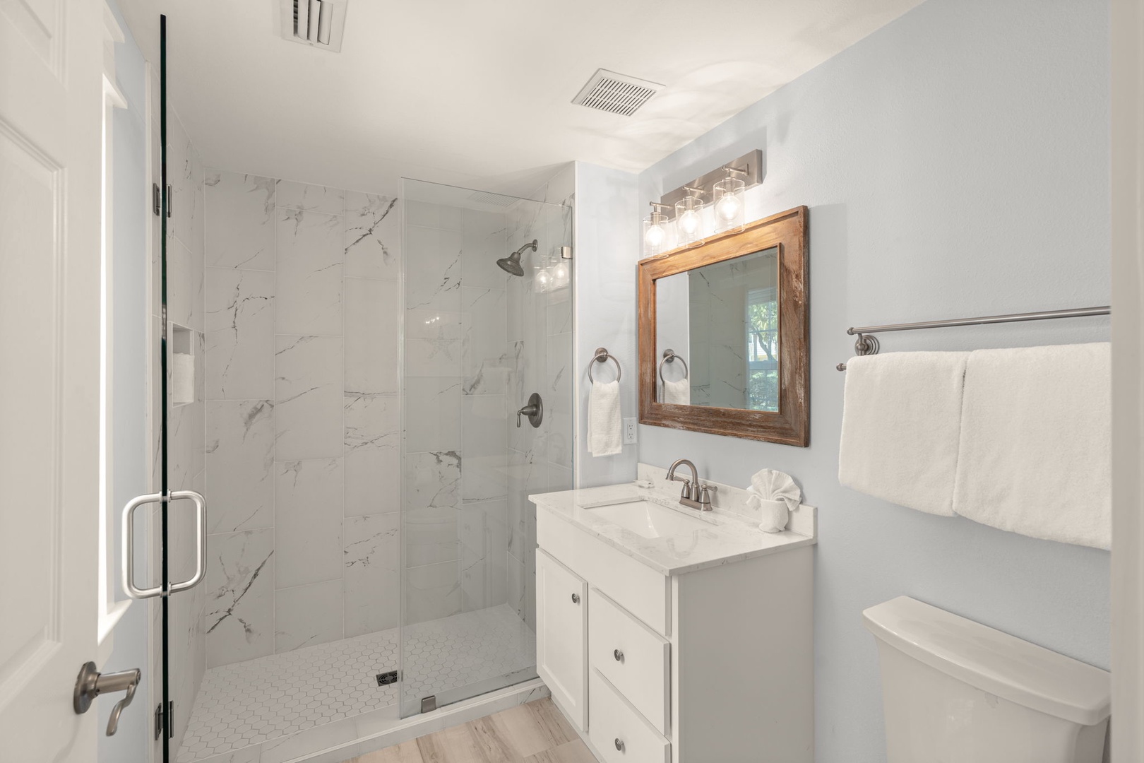 Your private bathroom features marble-look shower tiles, a wood-framed mirror above a white vanity, and fresh towels ready for your arrival.