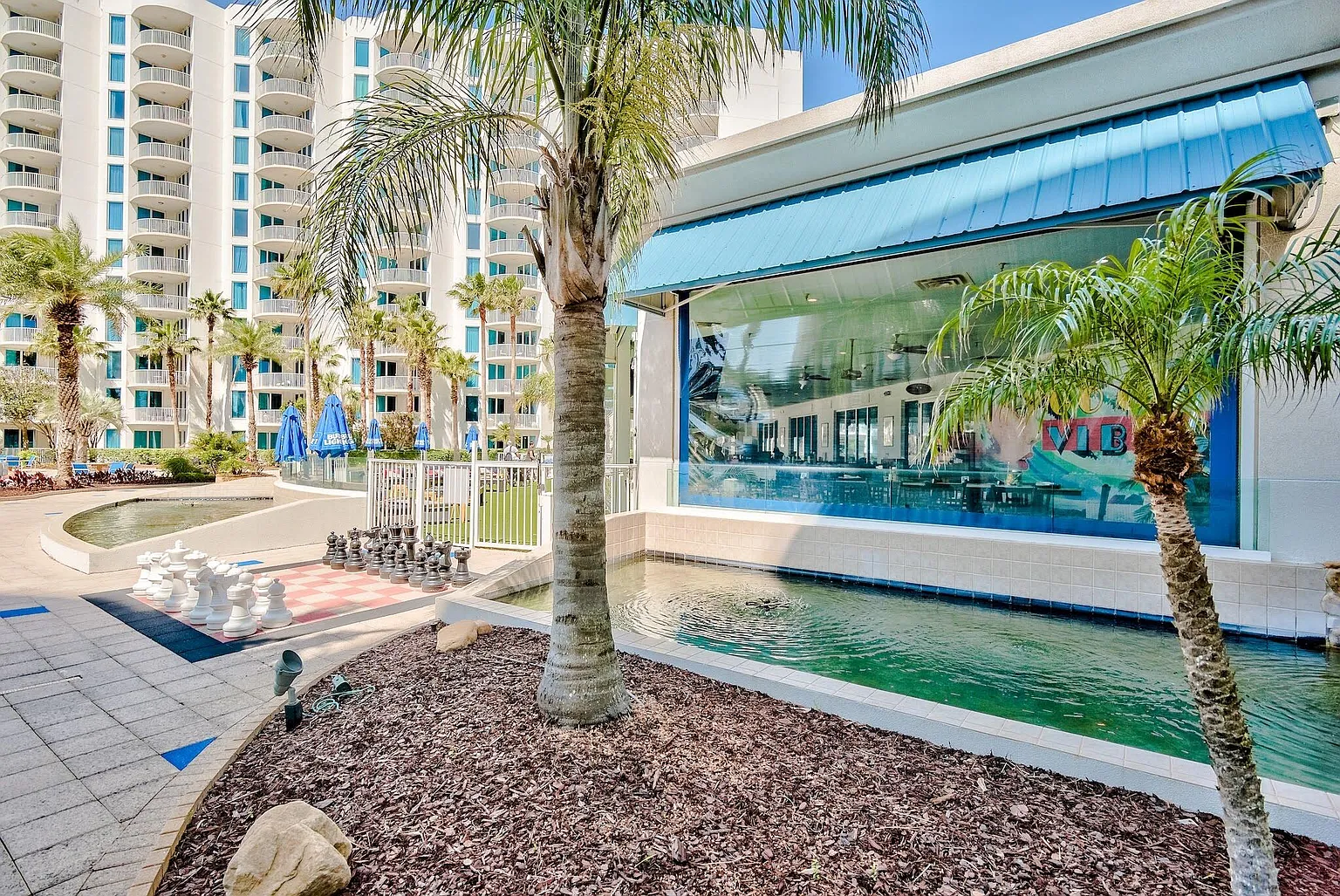 Large outdoor chess set and tropical landscaping with high-rise building in background.