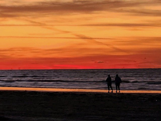 Golden sunset paints the sky over pristine beach where two silhouettes enjoy peaceful evening by the waves.