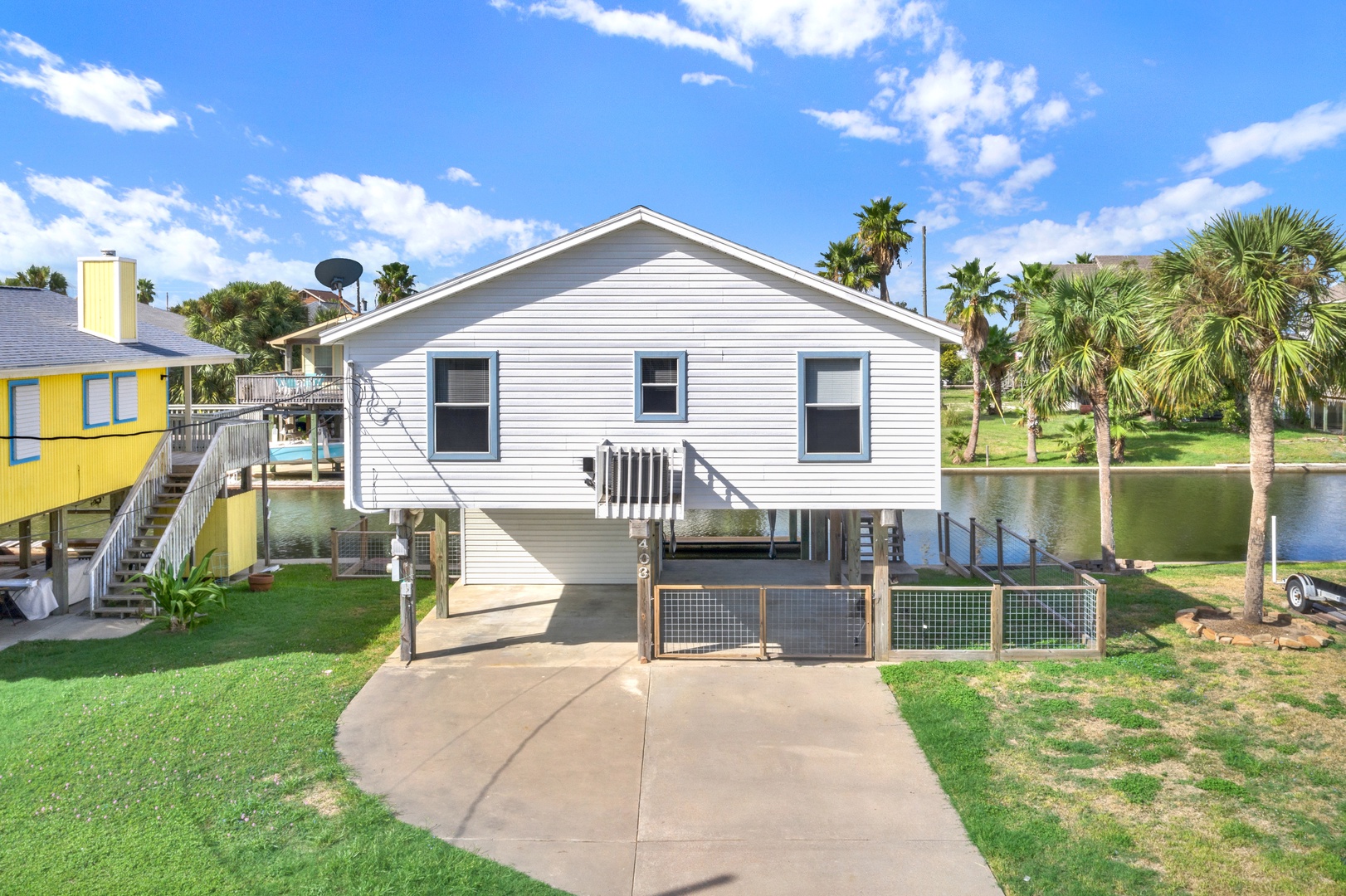 This charming coastal property sits elevated over tranquil waterfront grounds, surrounded by swaying palms and lush greenery under bright Texas skies—a peaceful escape with easy access to the shore.