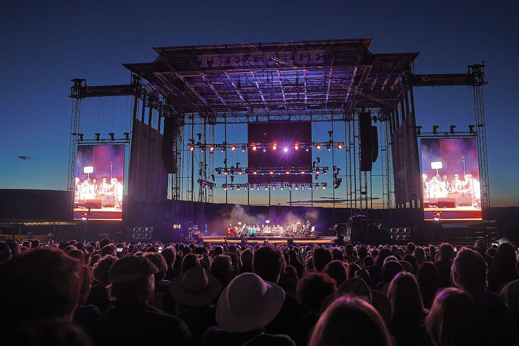 A stunning outdoor concert venue illuminated against the twilight sky, featuring a massive stage with dramatic lighting and large screens. This entertainment space hosts live performances in a scenic natural setting.