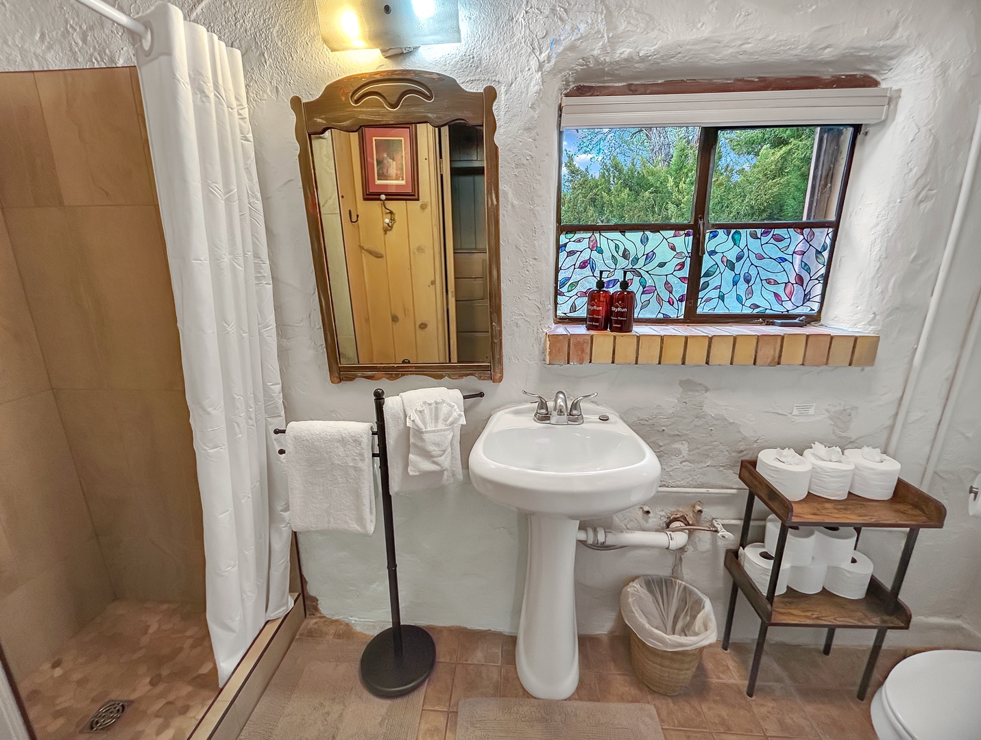 Your charming bathroom blends rustic character with modern comfort, featuring a pedestal sink and decorative mirror beneath a lovely garden window.