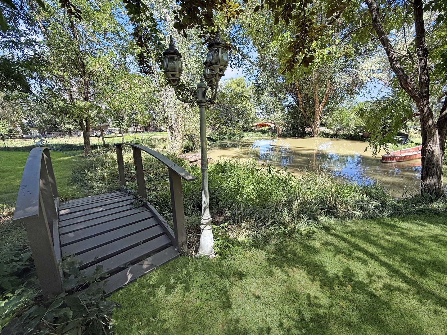A peaceful pond nestled among mature trees, framed by a charming footbridge and vintage lamppost, creates a tranquil setting perfect for morning walks or quiet reflection.