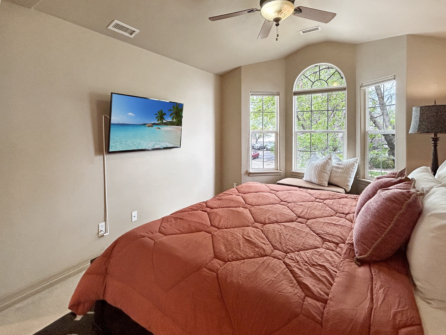 Your peaceful bedroom retreat features a plush bed, entertainment TV, gentle ceiling fan, and bright windows overlooking leafy surroundings.
