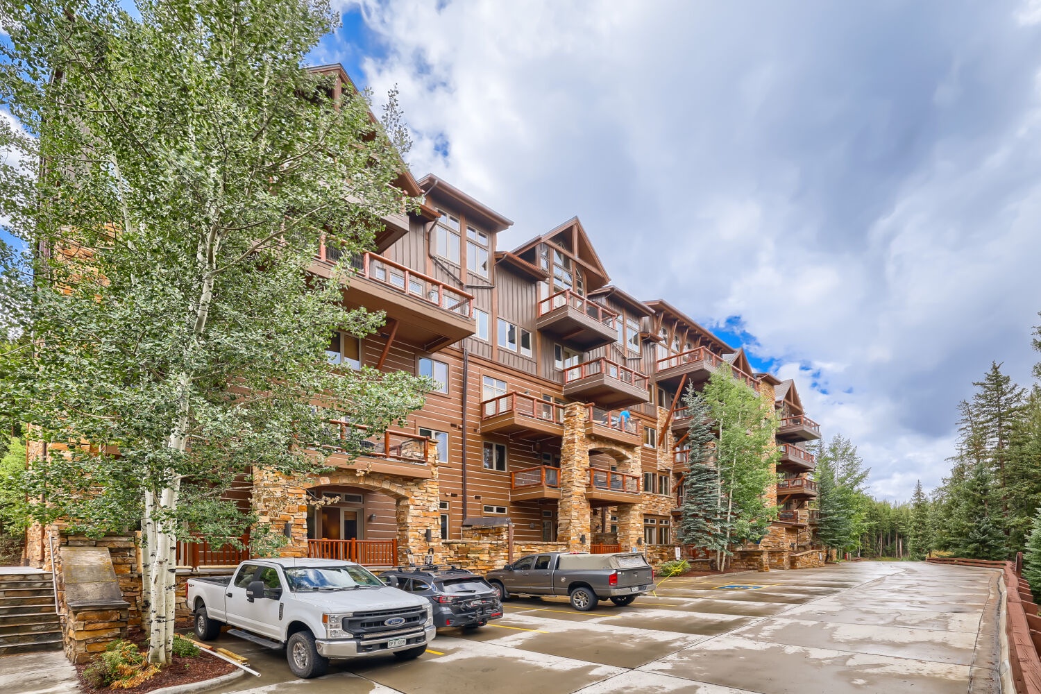This mountain lodge stands framed by towering aspens and evergreens, with convenient parking just outside. Stone and wood details capture the rustic alpine charm of the surrounding area.