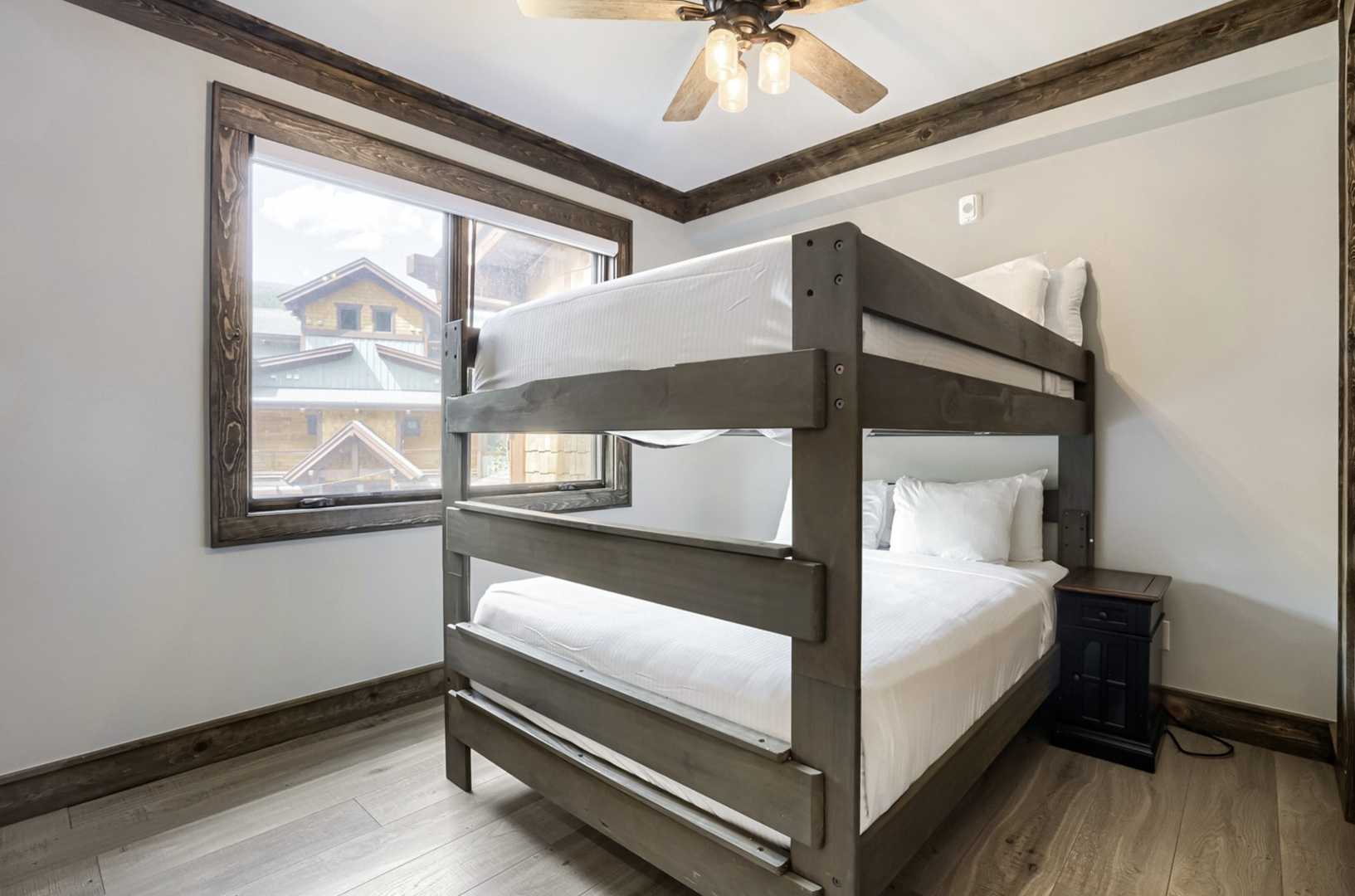 Cozy mountain bedroom with bunk beds and rustic charm, perfect for families or groups. Natural light pours through the window while wood beams and a ceiling fan add warmth overhead.