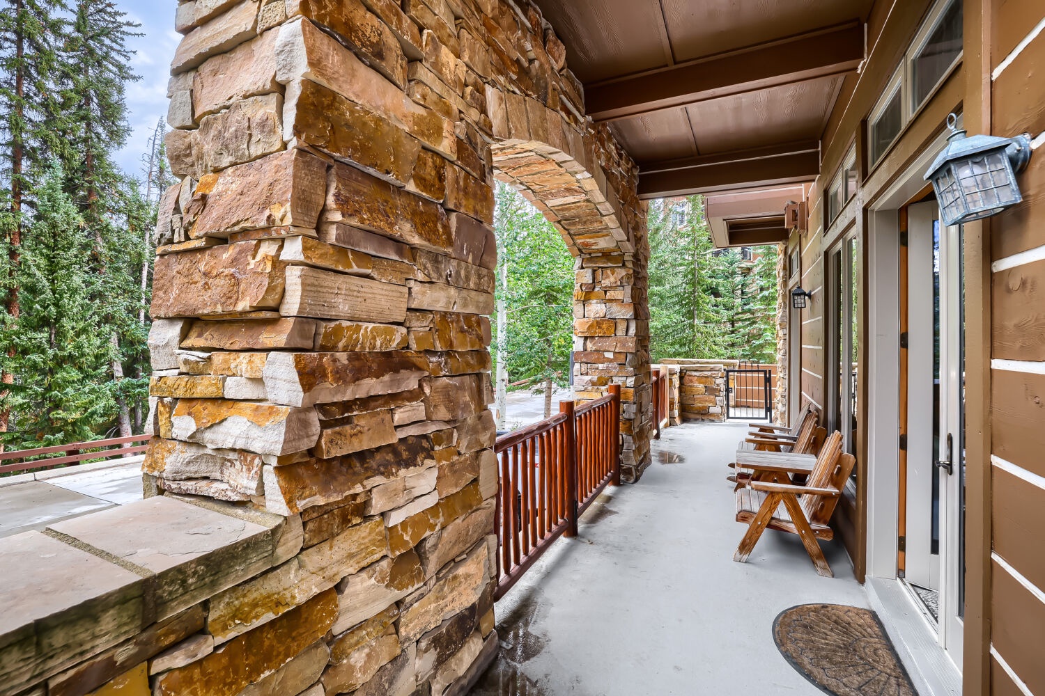 A spacious covered balcony featuring rustic stone pillars and wooden chairs invites you to relax surrounded by towering evergreens and fresh mountain air.