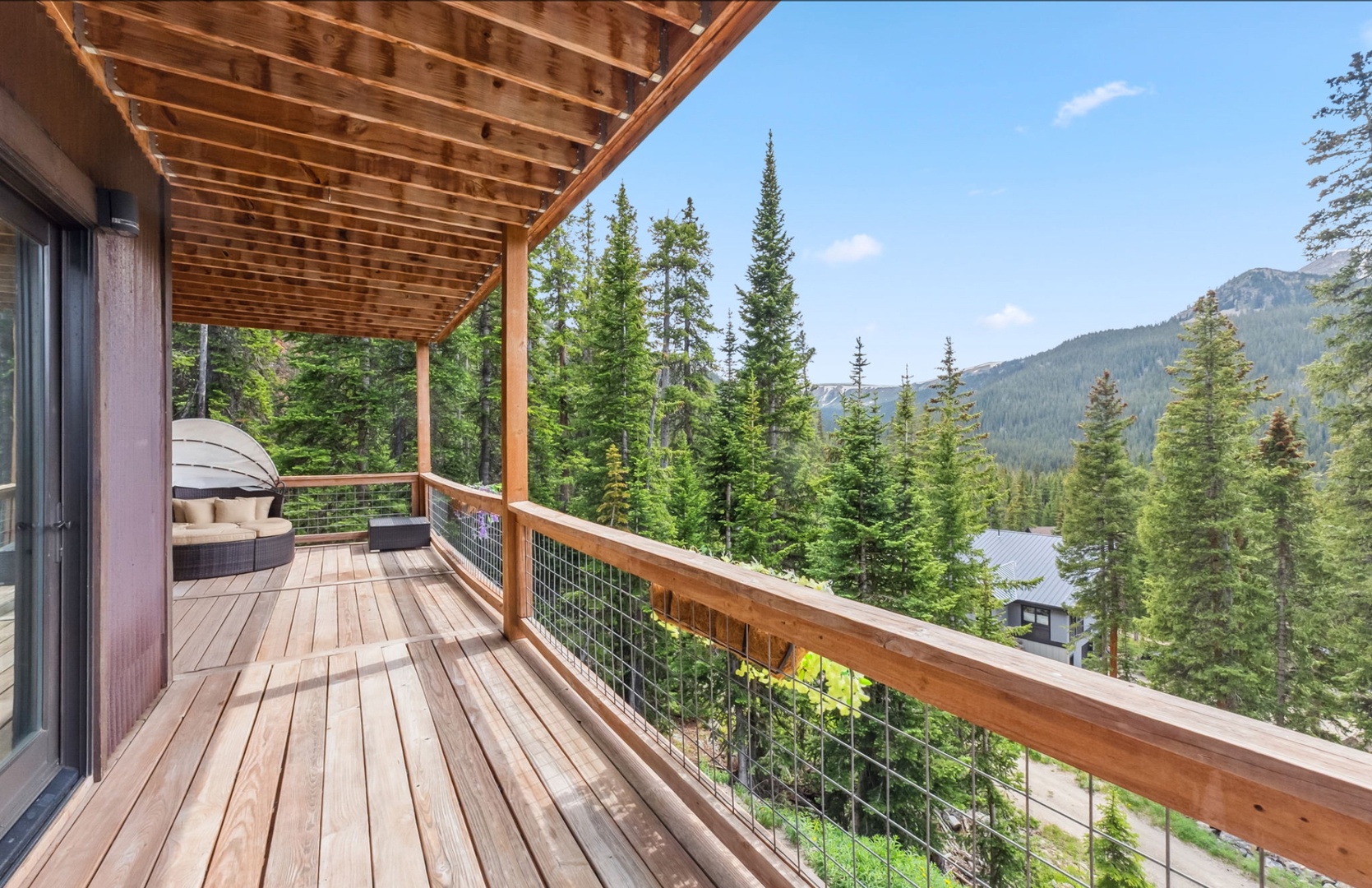 Expansive mountain views frame this covered deck with hot tub, surrounded by towering evergreens and dramatic alpine peaks on the horizon.