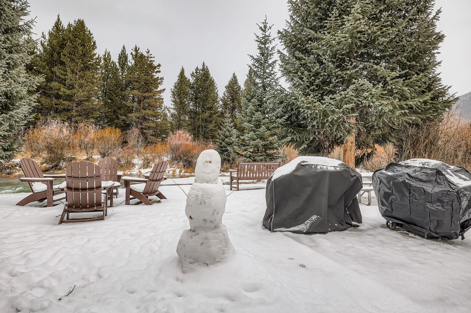 Snow-covered outdoor patio with Adirondack chairs and BBQ facilities, surrounded by evergreen trees in a peaceful winter setting