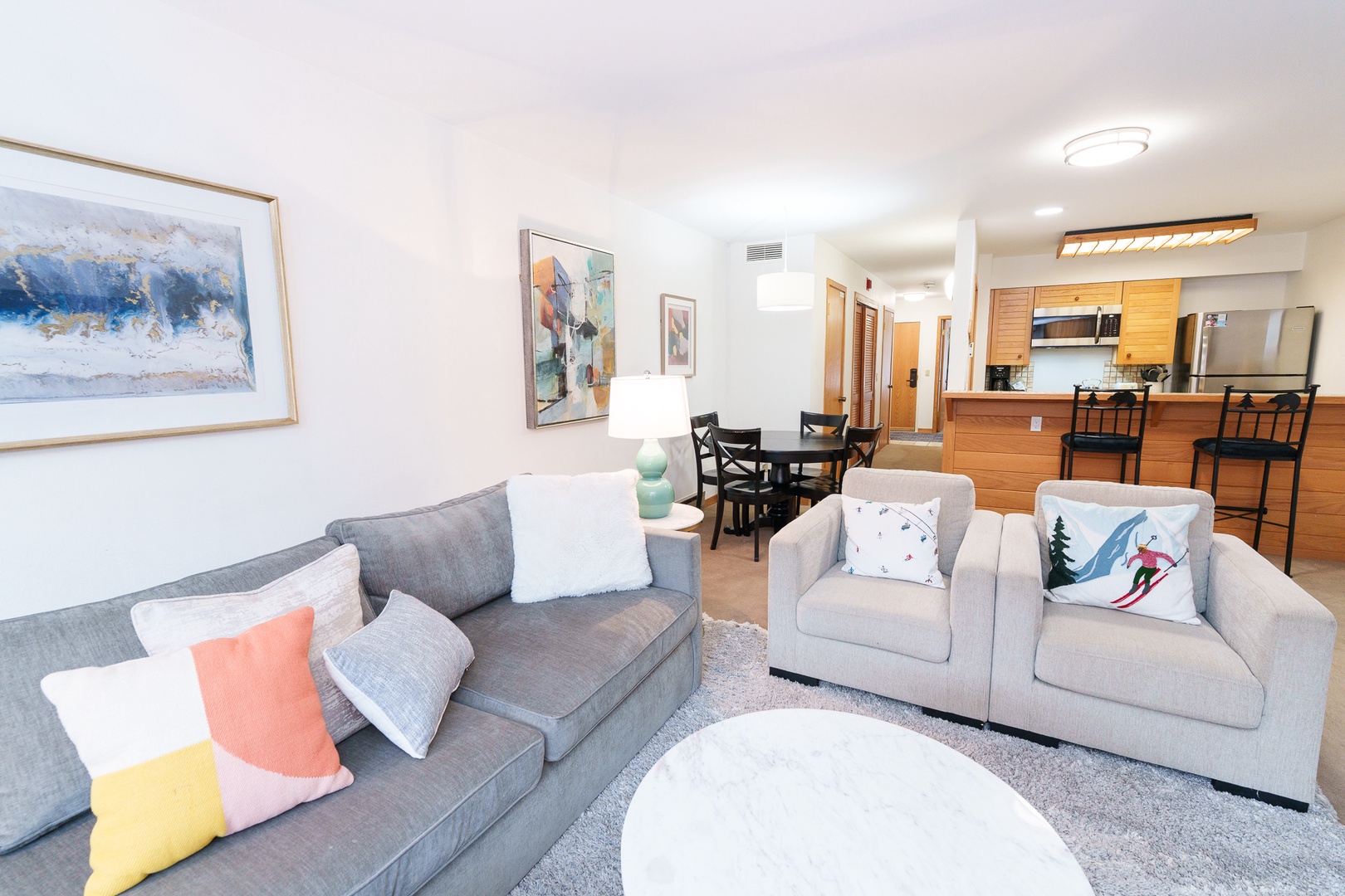 Relax in your spacious living area featuring plush gray sofas, colorful pillows, and thoughtful artwork, flowing seamlessly into your fully-equipped kitchen.