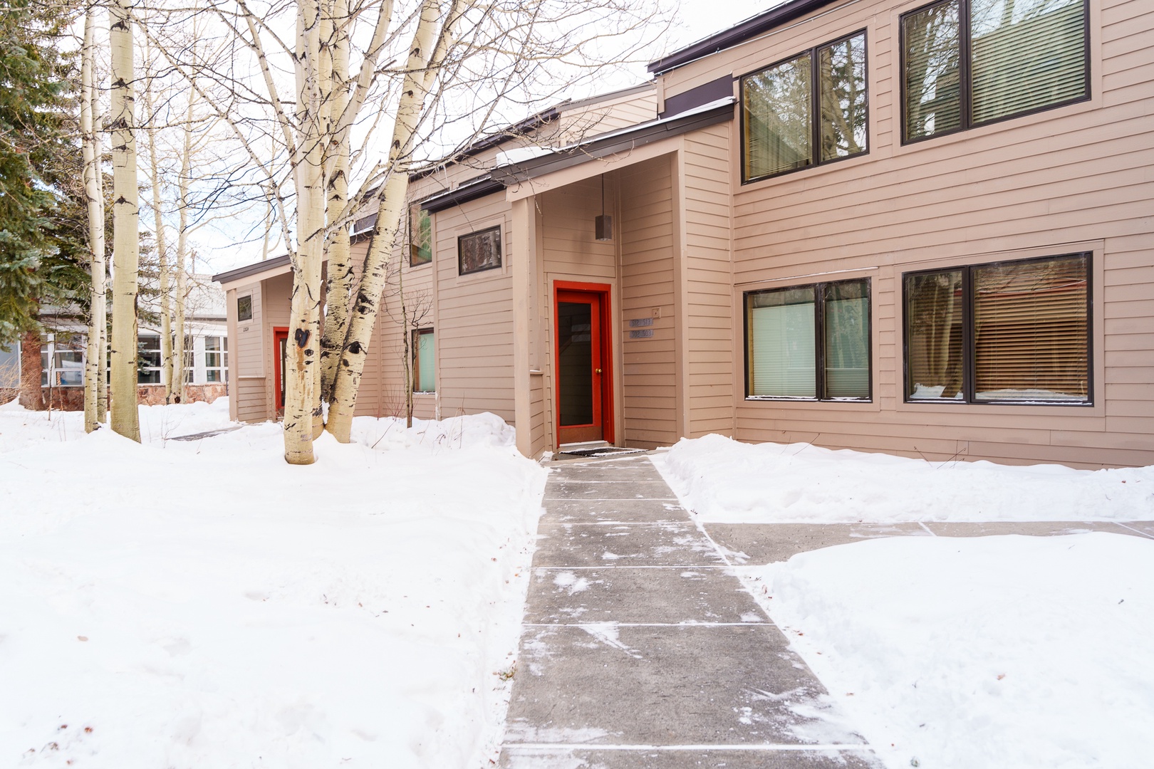Modern two-story property featuring warm wood siding and distinctive red entrance doors, set among snow-covered grounds and mature aspen trees.