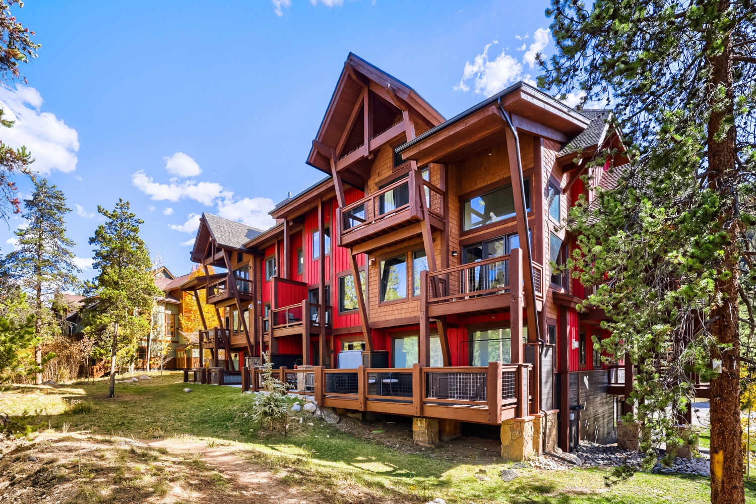 A striking red and wood mountain lodge nestled among towering pines, featuring dramatic peaked rooflines and expansive multi-level decks that invite outdoor relaxation in a pristine alpine setting.