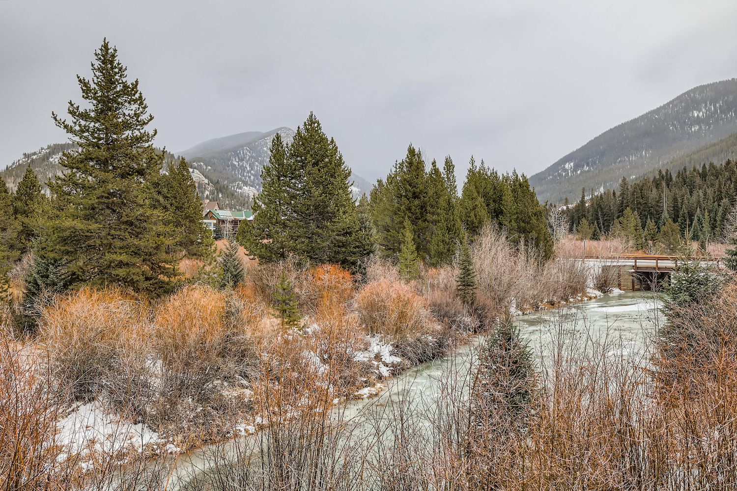 Mountain valley landscape with scattered snow and evergreen forest, surrounded by snow-capped peaks and winter terrain.