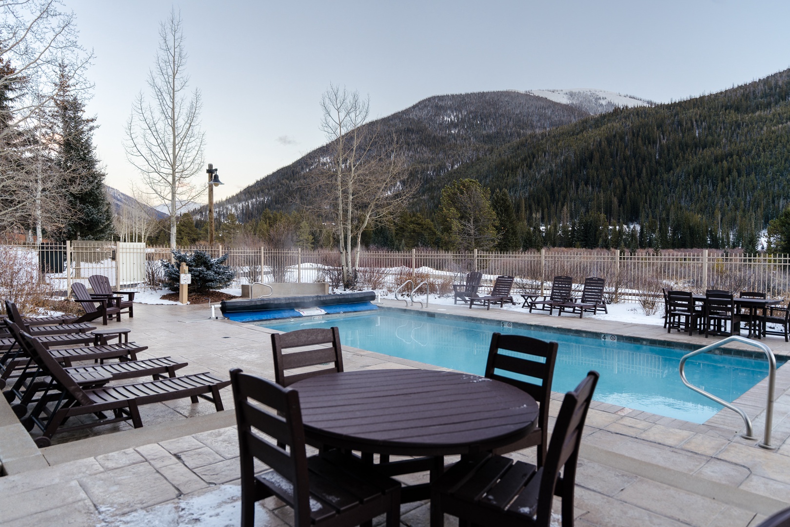Outdoor swimming pool area surrounded by snow-covered mountains and winter scenery, featuring poolside seating and dining furniture.