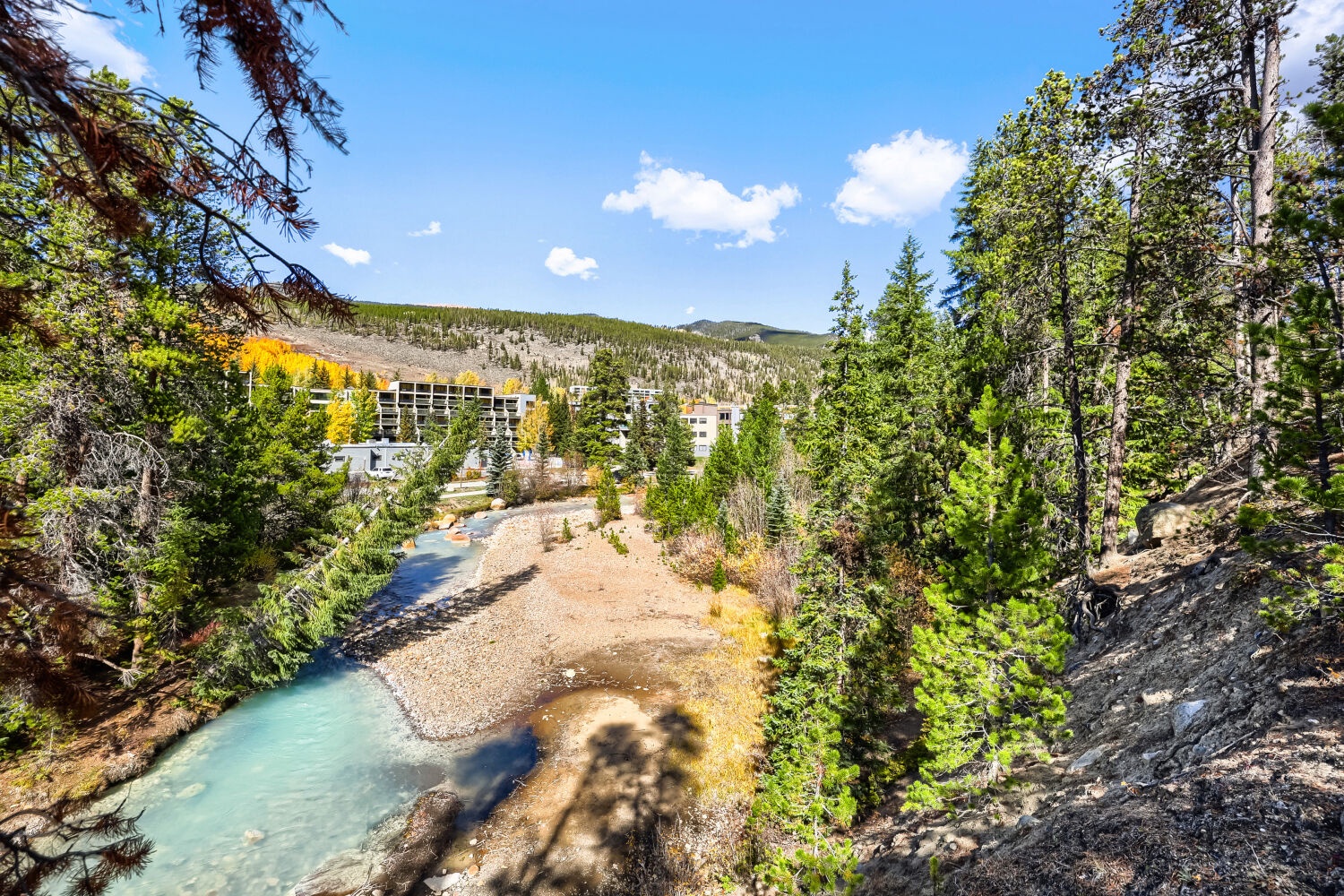 A pristine mountain river winds through evergreen forests and rugged terrain, with the property nestled into the scenic landscape beyond. Perfect for nature lovers seeking alpine tranquility.