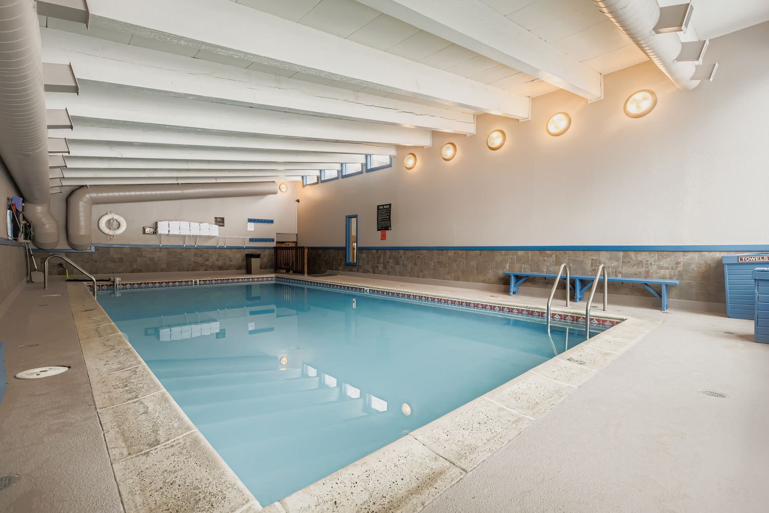 Community indoor pool with modern architectural beams and warm ambient lighting creating the perfect atmosphere for a refreshing swim.