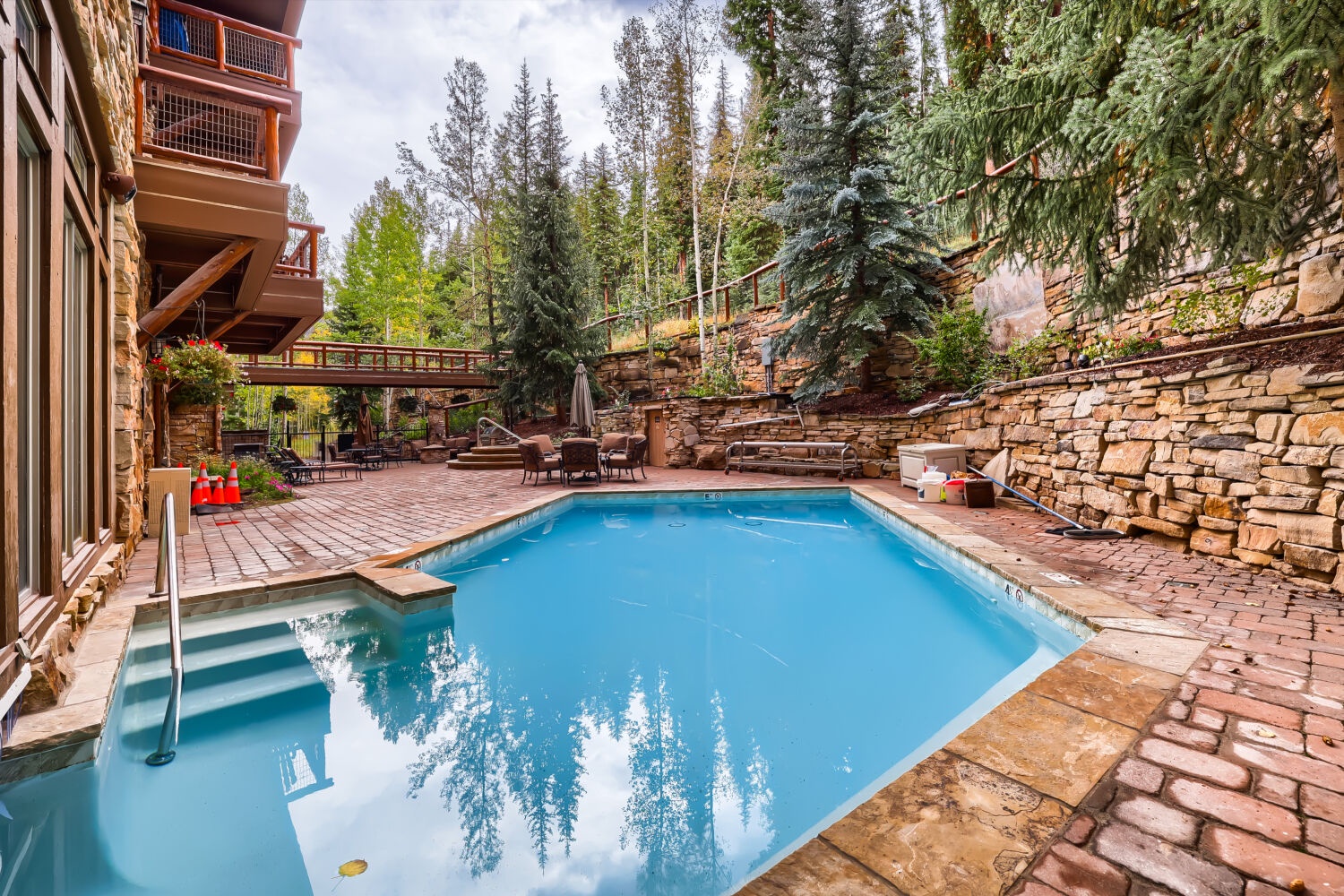 A serene outdoor pool nestled among evergreen trees and stone terraces invites you to unwind in nature's embrace, surrounded by rustic elegance and mountain tranquility.
