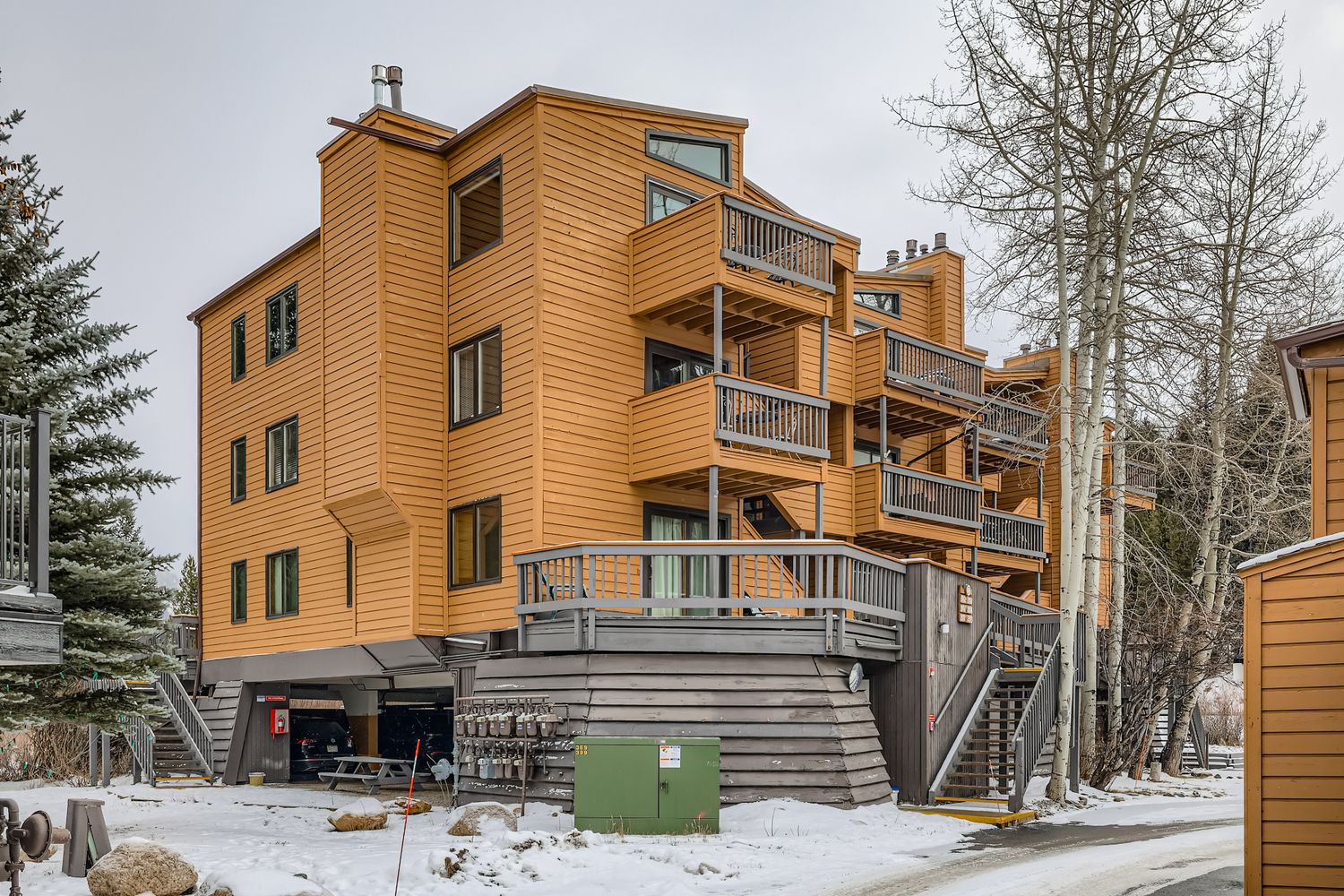Modern wood-sided property building with multiple balconies and exterior stairway access in a winter mountain setting.