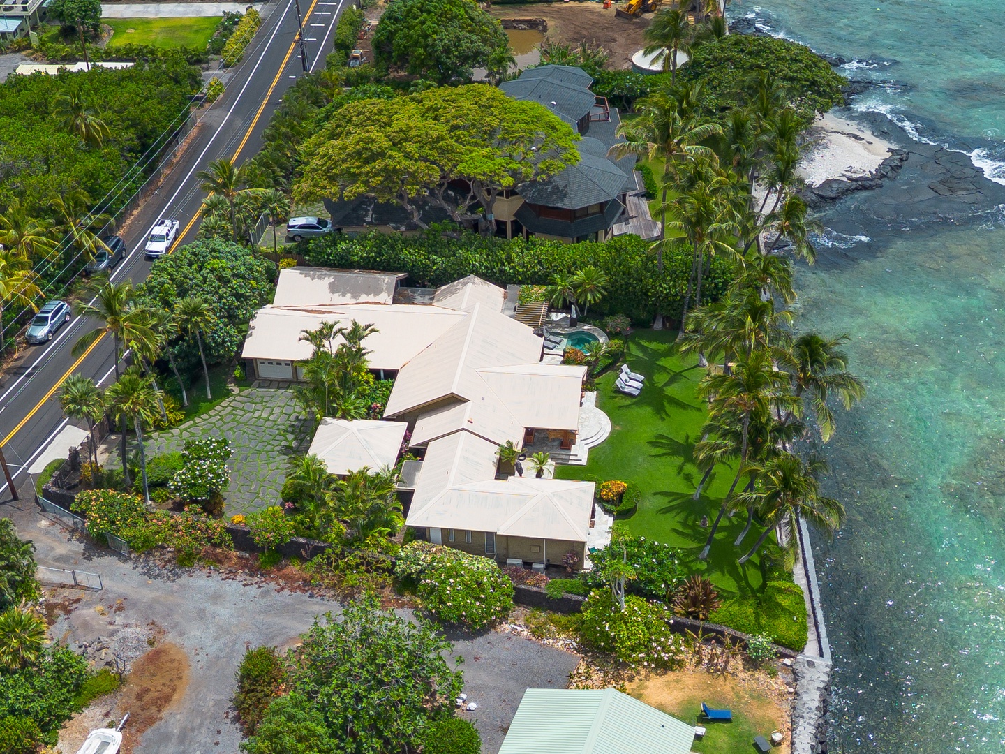 Aerial view of this stunning beachfront property nestled among lush tropical palms and greenery, with nearby ocean access and serene coastal surroundings along a quiet island road.