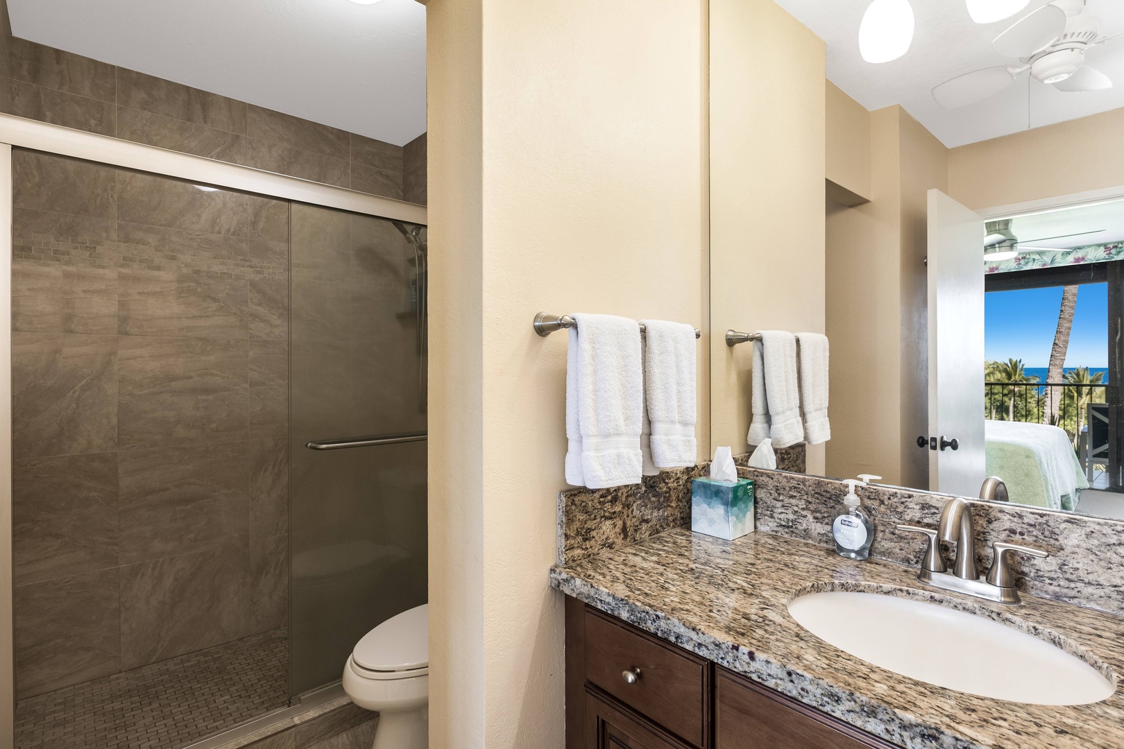Step into your private bathroom featuring a spacious walk-in shower and sleek granite countertops, with fluffy towels ready for your arrival. Catch glimpses of ocean blue through the window as you freshen up.
