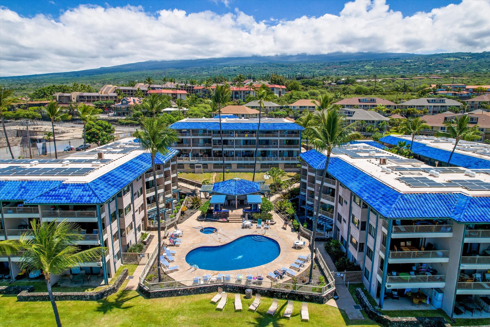 A resort-style property nestled on Hawaii's Big Island, Kona Reef features multi-level buildings with distinctive blue roofs surrounding a central pool courtyard, framed by palm trees with mountain and ocean views in the distance.