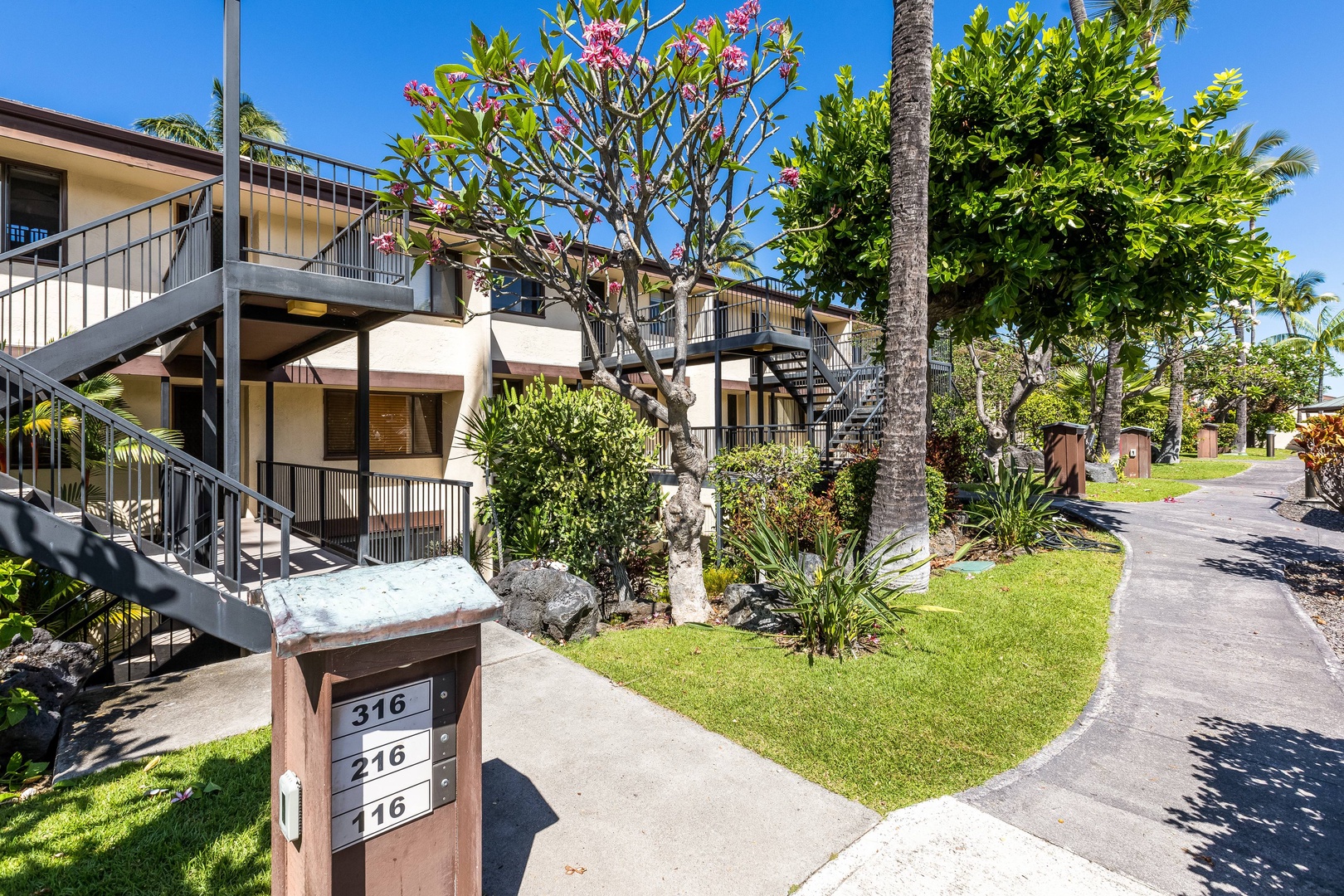 The property exterior welcomes guests with tropical landscaping, palm trees, and multi-level units arranged along a peaceful garden walkway in a sunny island setting.