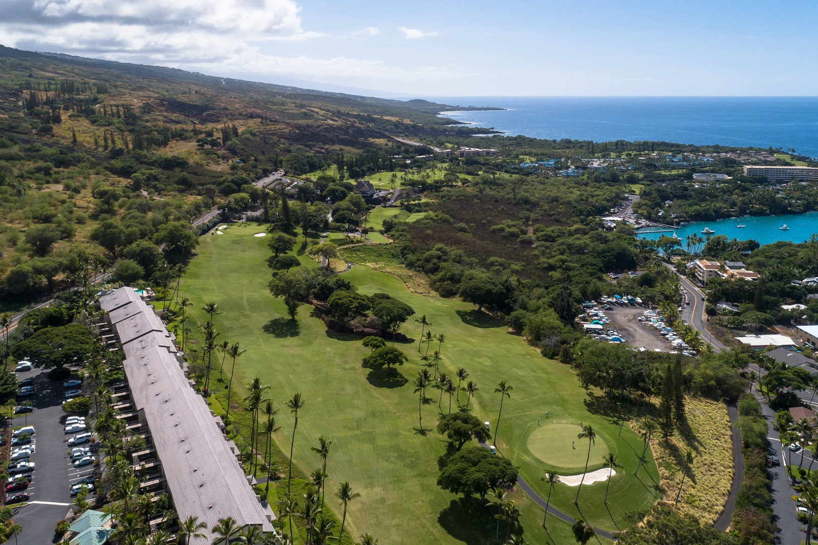 Aerial view of a coastal golf resort nestled between lush fairways and turquoise bay waters, with mountain slopes rising in the distance.