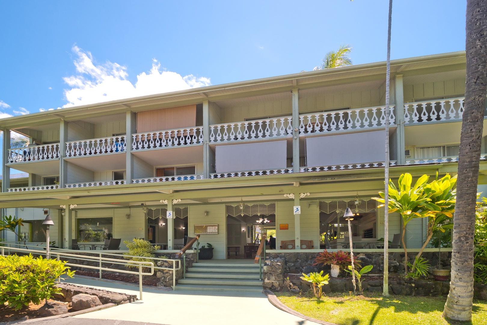 A charming two-story property welcomes guests with ornate balcony railings, lush tropical landscaping, and easy accessible entrance in a peaceful island setting.