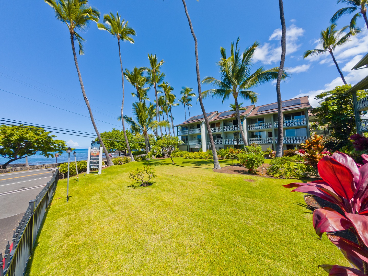 A coastal property features lush lawns and swaying palms between oceanfront and roadway, with tropical landscaping framing the blue-balconied buildings.