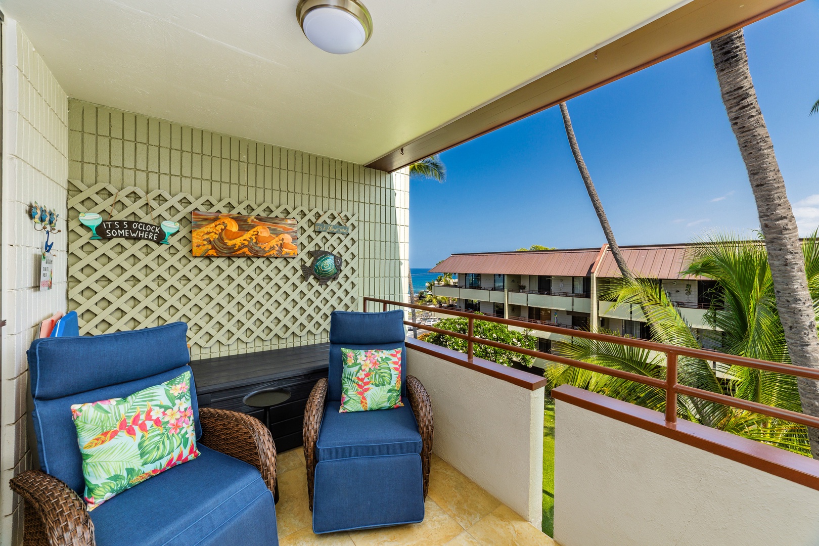 Your covered balcony is ready for morning coffee or evening relaxation, with comfy seating, cheerful tropical decor, and a lovely view of swaying palms and the sparkling ocean beyond.