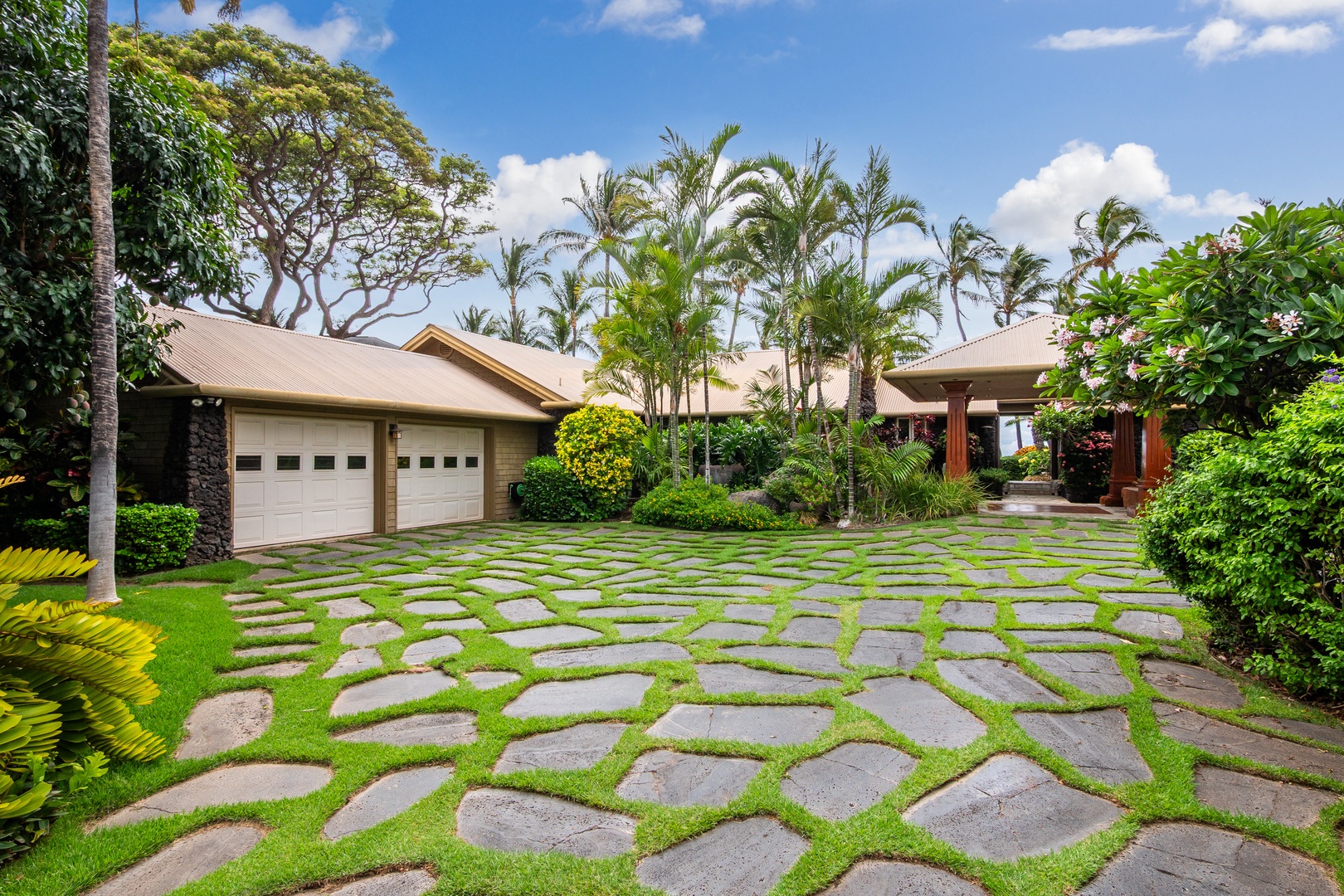 A tropical retreat framed by swaying palms and lush gardens, featuring a distinctive stone-paved driveway with grass accents leading to a contemporary island-style home.