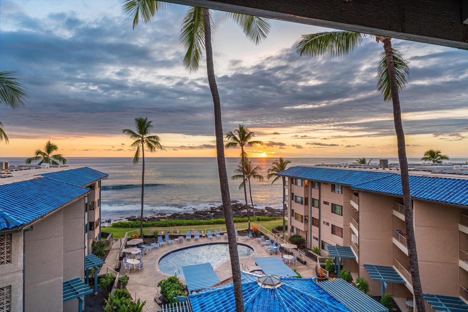 An oceanfront resort framed by swaying palms at sunset, where beachside buildings surround a tranquil pool deck overlooking the Pacific horizon.