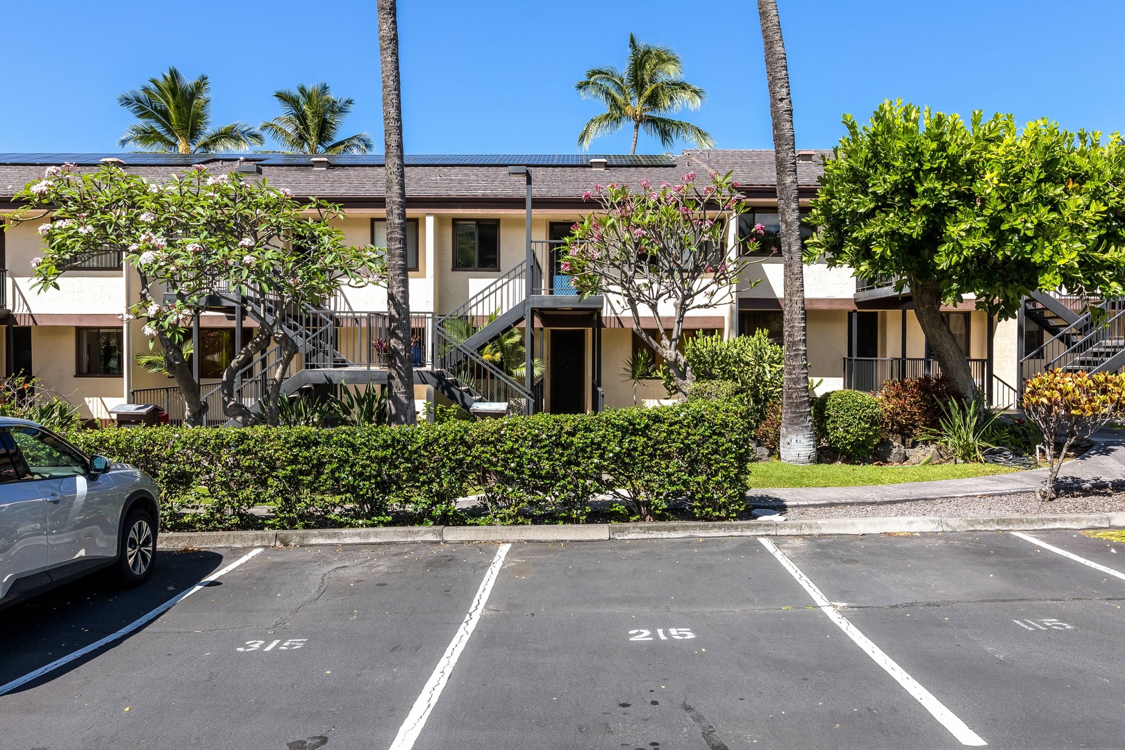 A two-story property building surrounded by lush tropical landscaping, including palm trees and vibrant flowering plants, with designated parking spaces located in front of the entrance.