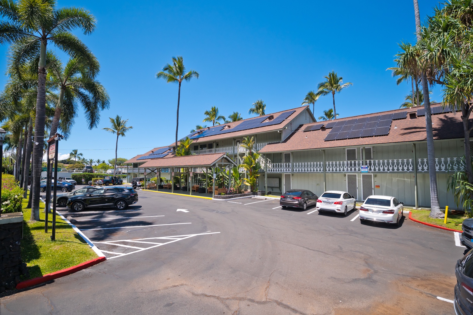 The property features multi-level buildings with solar panels, decorative balcony railings, and abundant parking surrounded by lush tropical palms beneath clear blue skies.