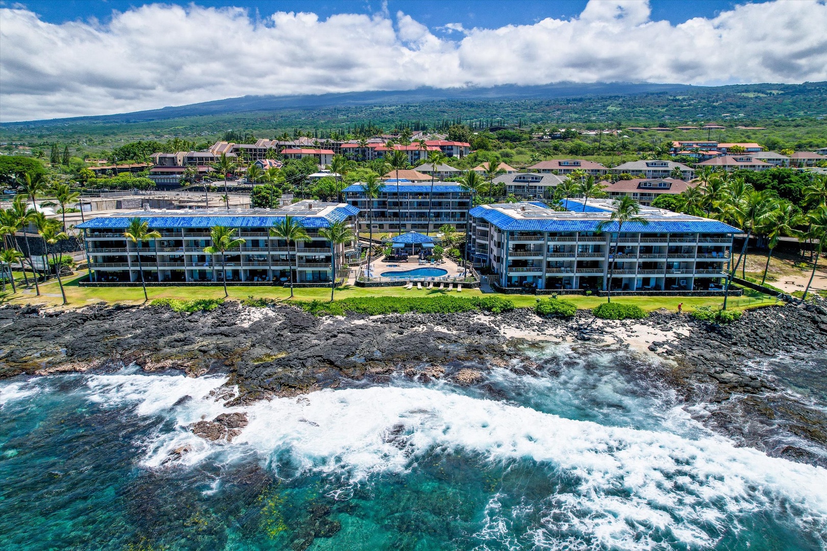 Oceanfront Kona Reef condo complex set on the Kona Coast shoreline with lush tropical grounds, swimming pool, and mountain backdrop.