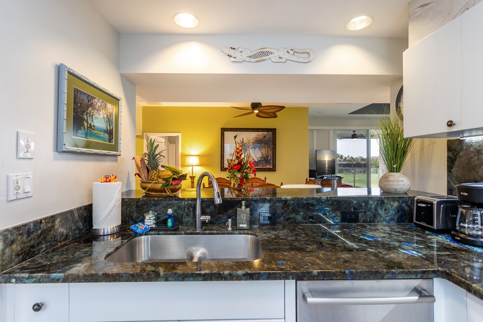 Your kitchen gleams with polished granite counters and stainless steel sink, opening to a bright dining and living space with tropical artwork and garden views beyond.