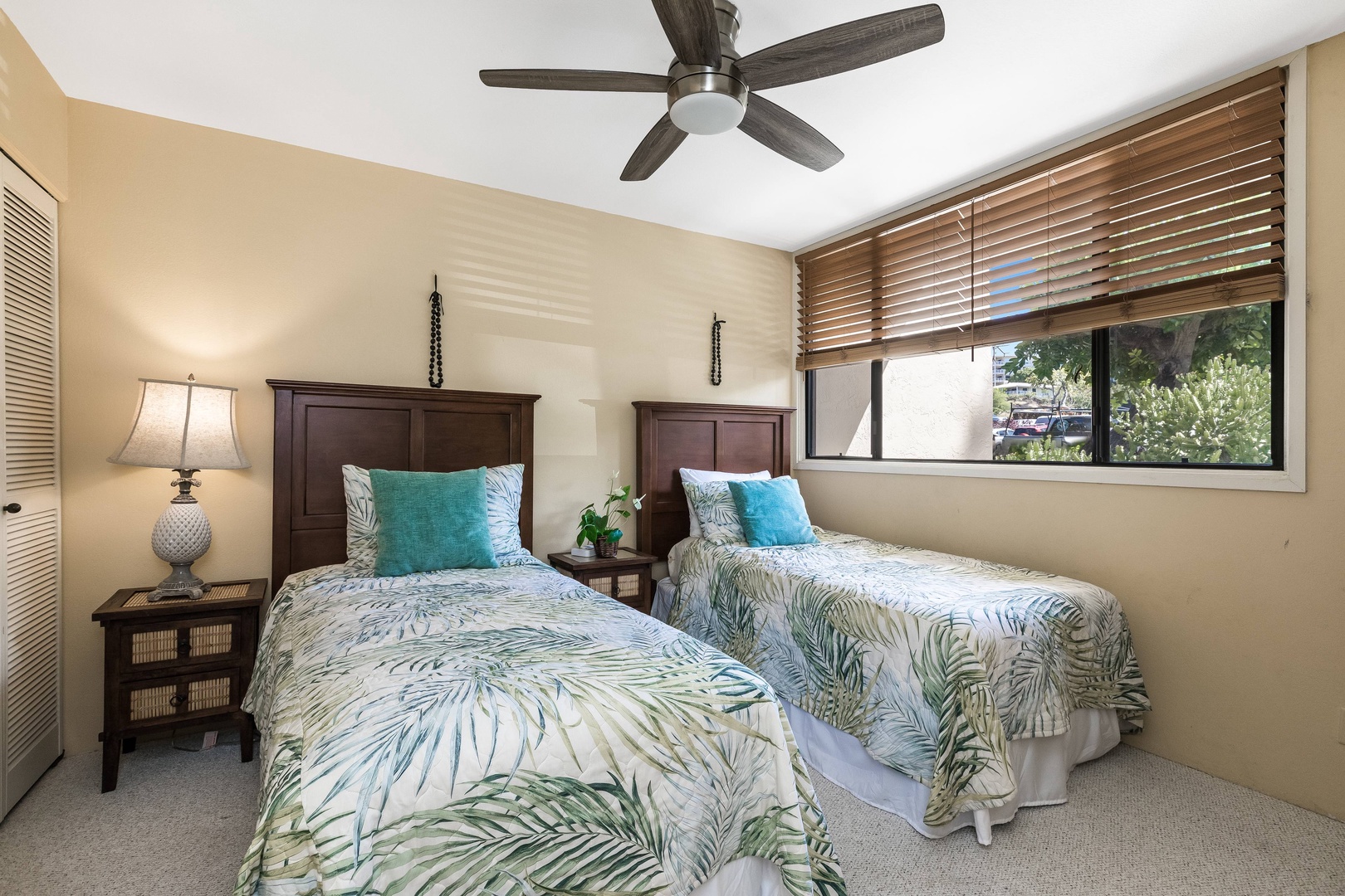 Your comfortable bedroom features two plush beds with tropical-inspired linens, a ceiling fan for cool breezes, warm natural light, and leafy views through generous windows that invite the outdoors in.