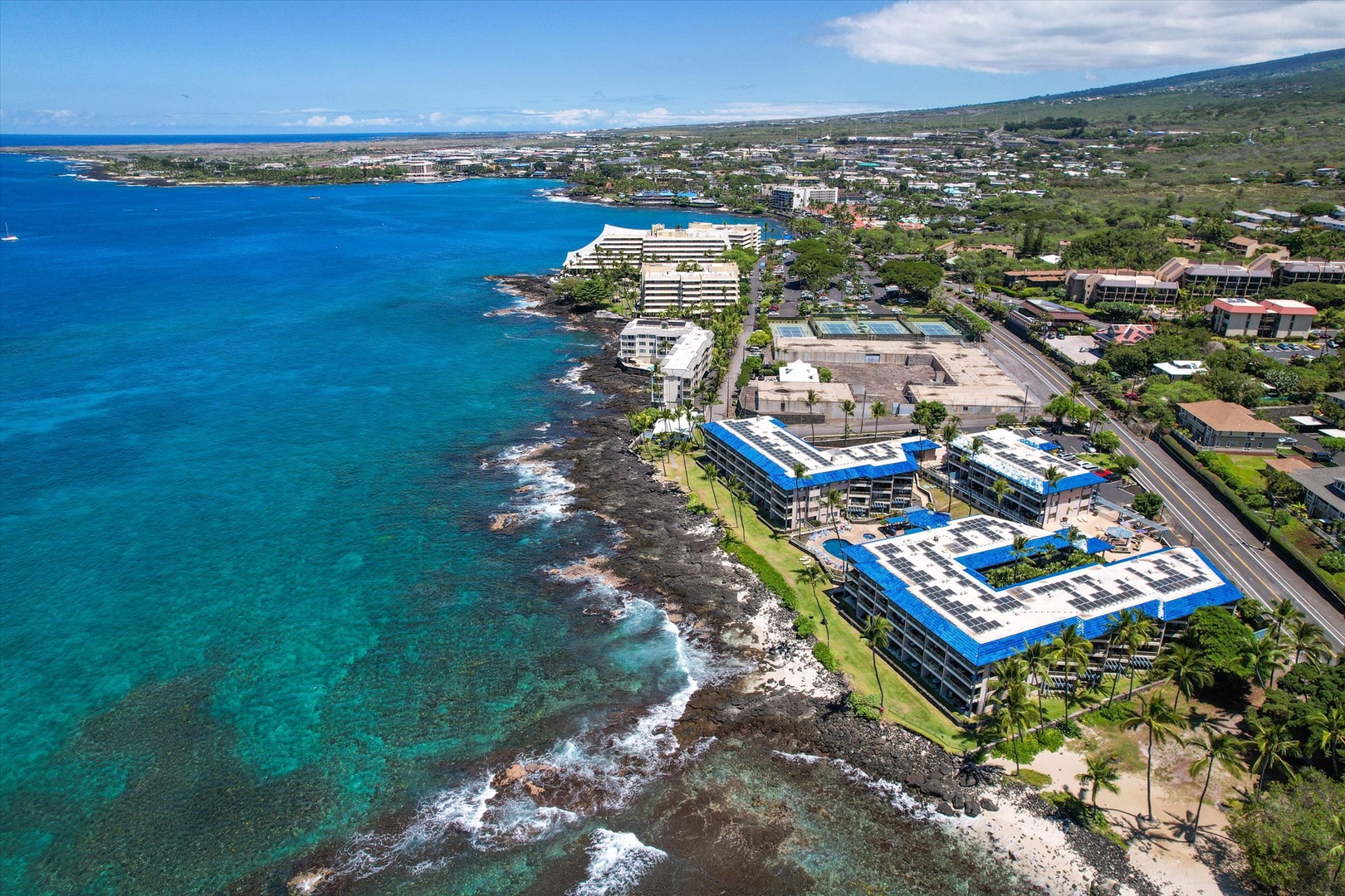 Kona Reef is situated along dramatic volcanic shoreline with turquoise waters, surrounded by tropical landscapes and neighboring resort developments in a serene island setting.
