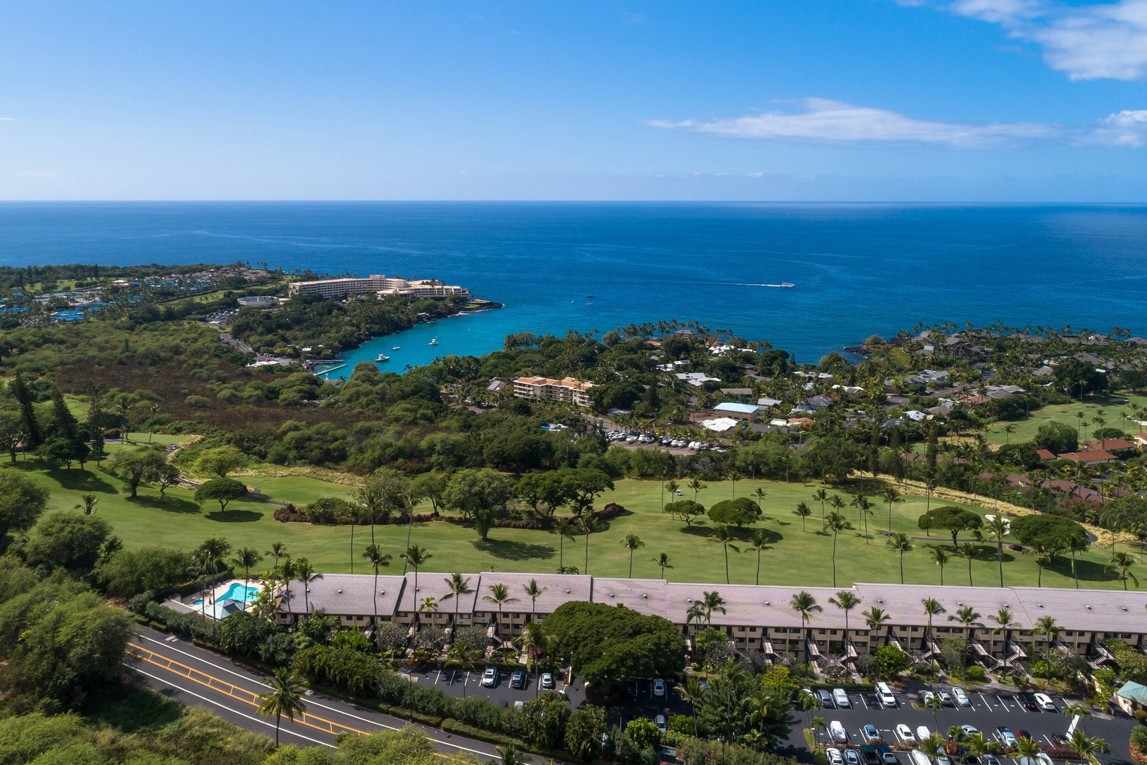 Stunning aerial view of a coastal resort property nestled between a championship golf course and brilliant turquoise bay waters, surrounded by tropical greenery and majestic ocean horizons.