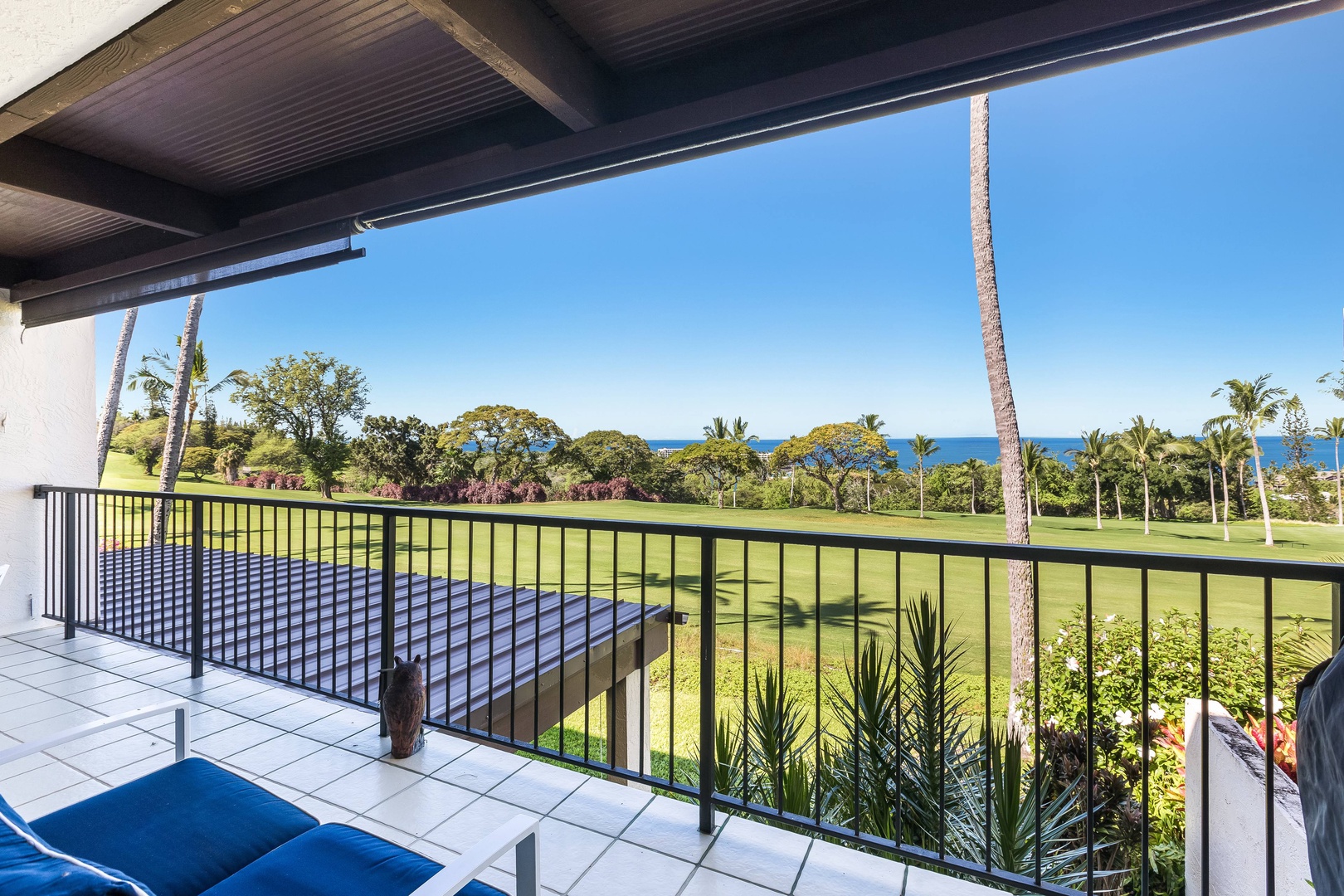 A vacation rental balcony overlooks a manicured golf course with palm trees and rolling greens extending toward the distant blue ocean under clear skies.