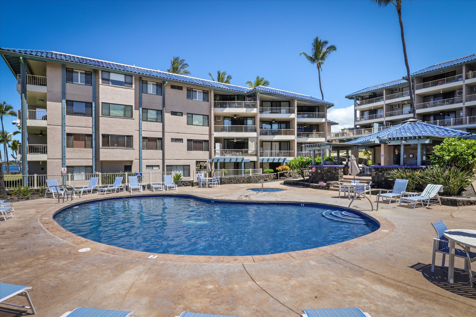 The property features a sparkling pool surrounded by spacious deck areas and tropical palms, set against multi-story buildings with private balconies under vibrant blue skies.