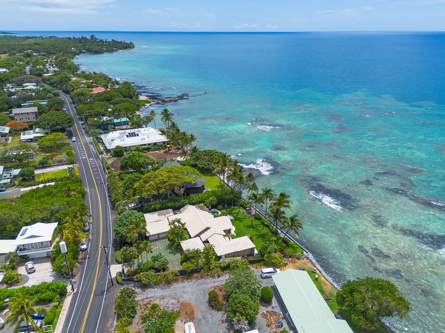 Aerial view of a coastal the Puako Beach Drive neighborhood with turquoise waters, lush tropical landscaping, and oceanfront homes lining a scenic coastal road.