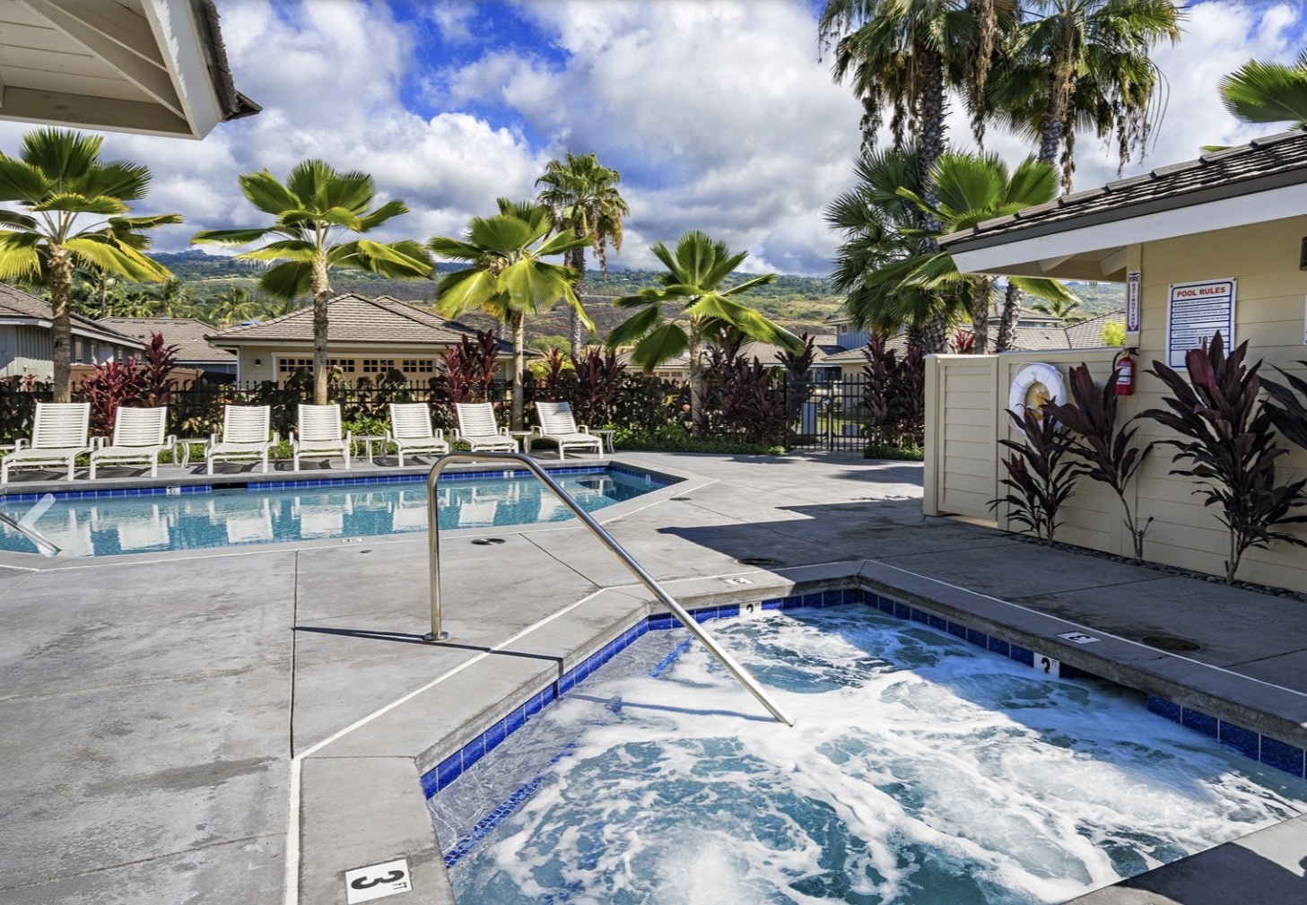 Tropical resort pool area with swimming pool, hot tub, and loungers surrounded by palm trees and mountain views.