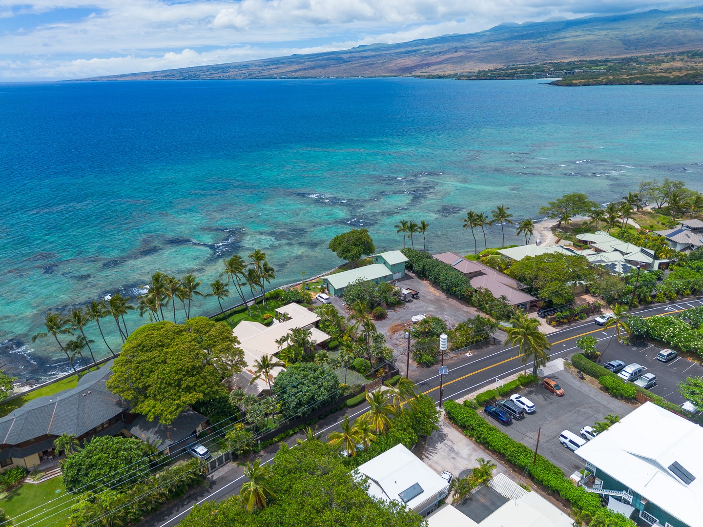 Aerial view of oceanfront property nestled along turquoise Hawaiian waters, with lush tropical landscaping, palm trees, and mountain backdrop.