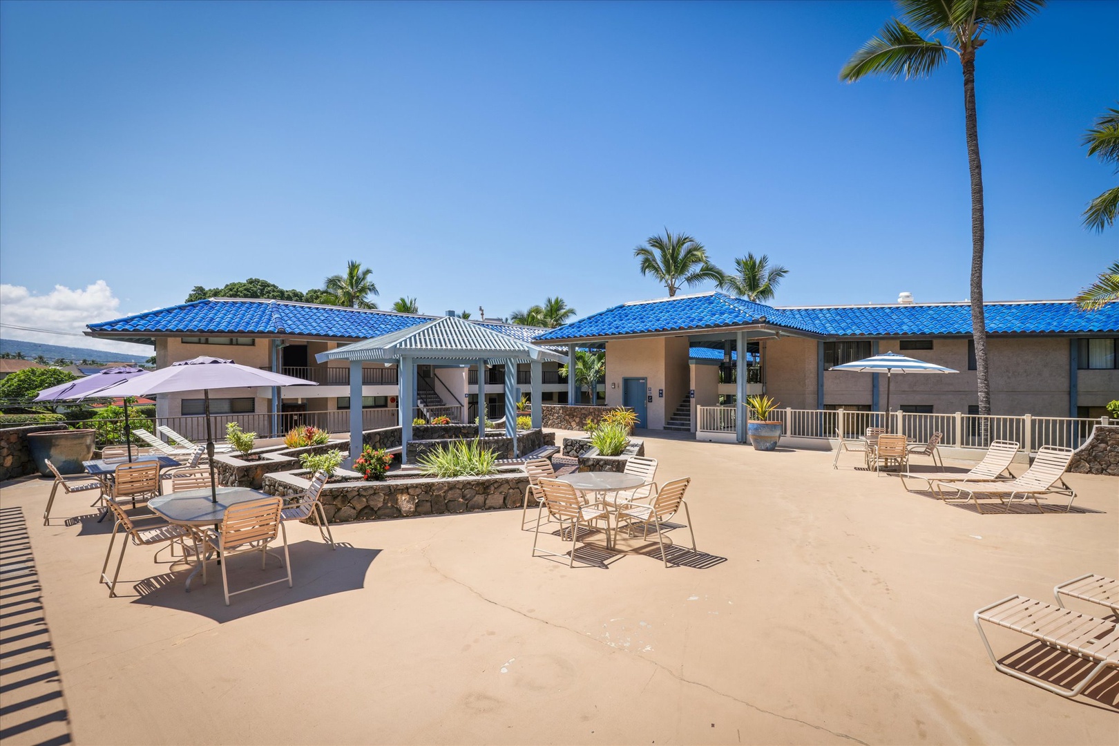 The property features tropical blue-roofed buildings surrounding spacious pool deck areas with lounge chairs, dining tables, and palm trees swaying in the breeze.