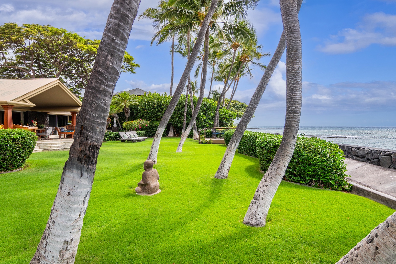 The property features a lush oceanfront lawn framed by swaying palms, with a stone Buddha statue nestled in the grass and direct access to the shoreline.
