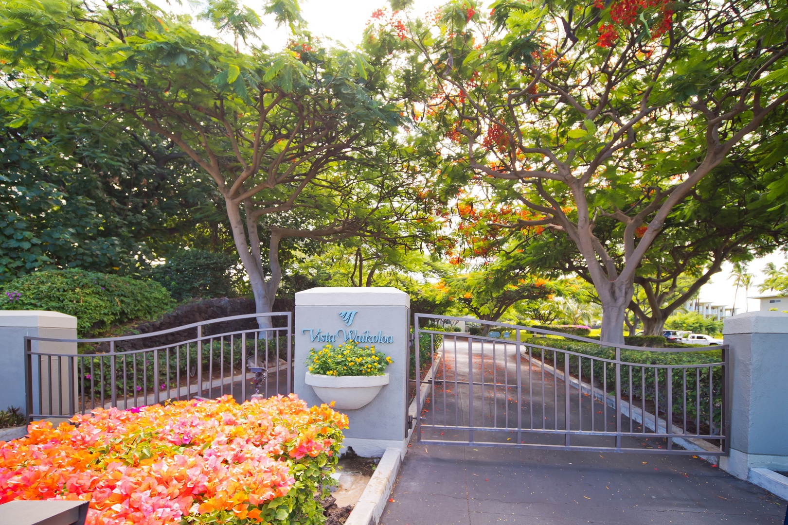 Property entrance marker with landscaped gardens and metal gates beneath flowering trees. The Vista Waikoloa sign welcomes arrivals to this tropical vacation community.