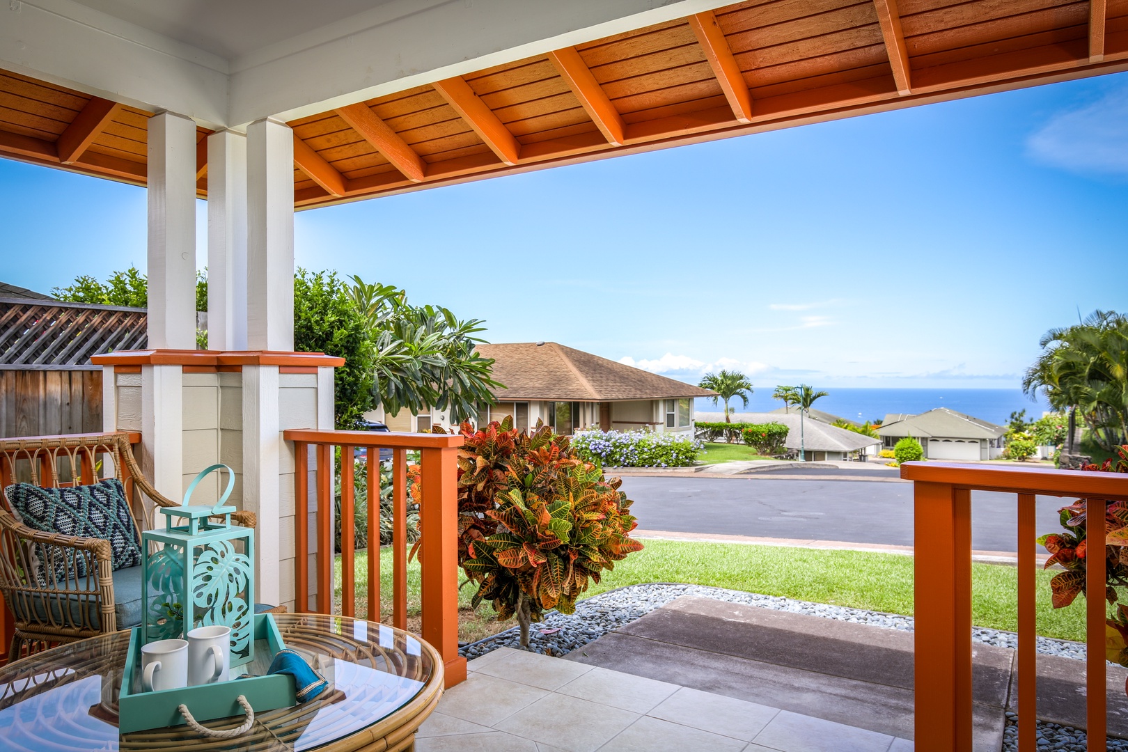 A covered lanai frames sweeping views of a tranquil residential neighborhood, with distant ocean vistas beyond palm-dotted homes. The property entrance is accented by lush tropical landscaping and clean, modern woodwork.