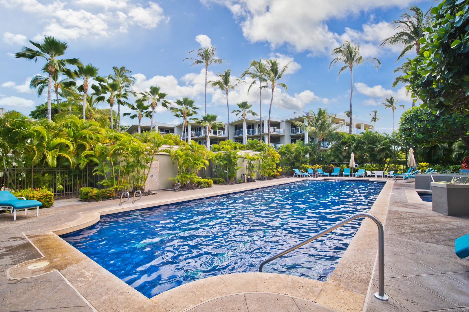 The sparkling pool awaits beneath swaying palms, with turquoise loungers and tropical landscaping creating a perfect spot to unwind under sunny skies. The modern property building stands peacefully in the background.