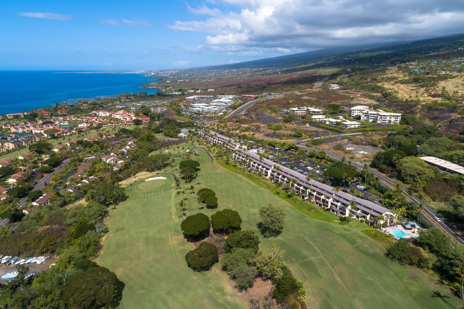 Aerial view of a coastal resort community featuring a golf course, residential buildings, and stunning ocean vistas stretching to the horizon along Hawaii's scenic coastline.
